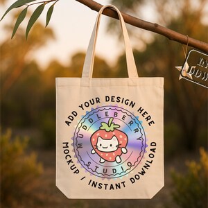May include: A cream-coloured tote bag featuring a strawberry cat design. The bag has handles and text that reads "Add Your Design Here" and "Mockup / Instant Download". The background is blurred with natural light.