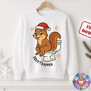 Könnte beinhalten: Weißes Sweatshirt mit einem Cartoon-Eichhörnchen, das einen Weihnachtsmannhut trägt und auf einer Toilette sitzt. Der Text "Nut Crapper" steht unter dem Eichhörnchen. Das Design enthält Sterne.