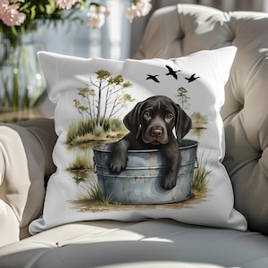 Könnte beinhalten: Weißes quadratisches Kissen mit einer Aquarellillustration eines braunen Welpen in einem Metalleimer. Der Hintergrund zeigt einen Teich, Bäume und Vögel. Das Kissen liegt auf einem cremefarbenen Sofa.