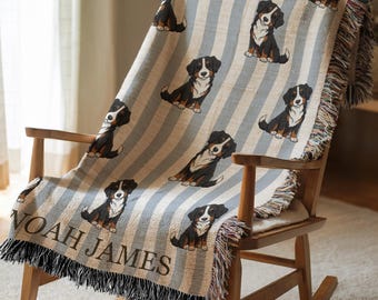 May include: A woven throw blanket draped over a wooden rocking chair. The blanket features a repeating pattern of Bernese Mountain Dog illustrations on a striped background of light blue and beige. The name "NOAH JAMES" is woven into the bottom of the blanket.