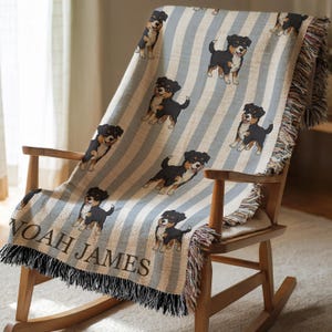 May include: A woven throw blanket featuring a repeating pattern of black, brown, and white dogs on a light blue and white striped background. The blanket is draped over a wooden rocking chair. The name "NOAH JAMES" is displayed.
