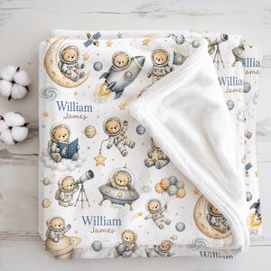 Op de afbeelding: Een witte fleece deken met een herhalend patroon van teddyberen in astronautenpakken, raketten, planeten, sterren en telescopen. De tekst William James is gedrukt in blauw en bruin schrift. De deken heeft een zachte, witte pluche onderkant.