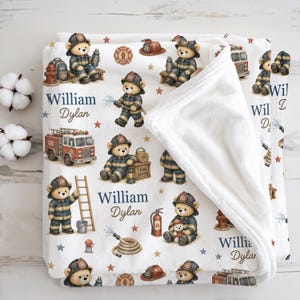 May include: A white fleece blanket featuring a repeating pattern of teddy bears dressed as firefighters, red fire trucks, fire hydrants, and hoses. The text William Dylan is printed in blue and brown script. The blanket has a soft white underside.