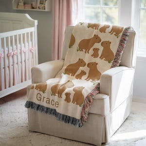 K&ouml;nnte beinhalten: Eine beige Decke mit einem sich wiederholenden Muster aus braunen Capybaras und ihren Jungen. Die Decke ist &uuml;ber einem wei&szlig;en Sessel in einem Kinderzimmer drapiert. Der Name "Grace" ist auf die Decke gedruckt.