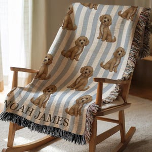 Op de afbeelding: Een geweven sprei met een herhalend patroon van bruine puppy's op een lichtblauwe en crèmekleurige gestreepte achtergrond. De sprei is gedrapeerd over een houten schommelstoel. De naam "NOAH JAMES" is in het zwart gedrukt in de linkerbenedenhoek, met een zwarte franje.