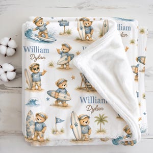 Puede incluir: Una manta de forro polar blanca con un patrón repetitivo de ositos de peluche surfeando y sosteniendo tablas de surf. El diseño incluye palmeras, olas, estrellas de mar y un letrero que dice "SURF'S UP". El nombre "William Dylan" está impreso en azul.