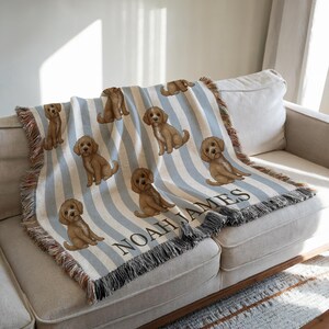 May include: A woven throw blanket featuring a repeating pattern of brown puppies on a light blue and white striped background. The blanket has a fringed edge and the name "NOAH JAMES" is displayed in black text.