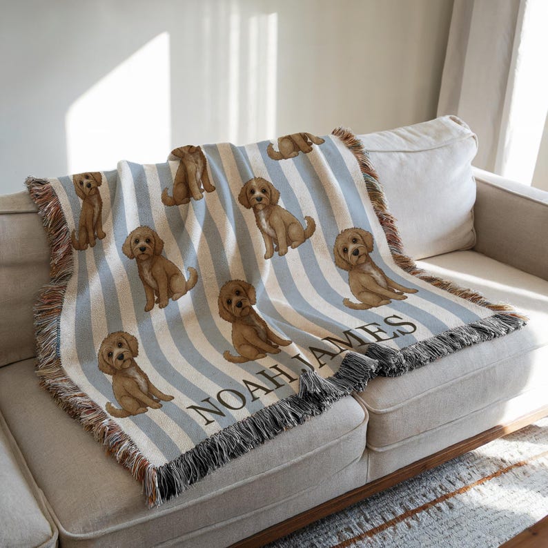 May include: A woven throw blanket featuring a repeating pattern of brown puppies on a light blue and white striped background. The blanket has a fringed edge and the name "NOAH JAMES" is displayed in black text.