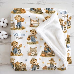 Puede incluir: Una manta de forro polar blanca con un patrón repetitivo de osos de peluche con petos, calabazas, linternas y letreros de madera con la etiqueta Pumpkin Patch. La manta incluye el texto personalizado William Dylan en fuentes azules y marrones.