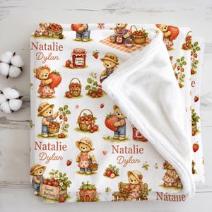May include: A white fleece blanket featuring a repeating pattern of teddy bears picking strawberries, carrying baskets of fruit, and jars of jam. The text Natalie Dylan is printed in a brown script font throughout the design on the soft fabric.