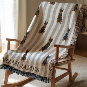 May include: A blue and white striped throw blanket with a repeating pattern of black and brown Doberman Pinscher dogs. The blanket is draped over a wooden rocking chair. The name "NOAH JAMES" is printed on the bottom edge.