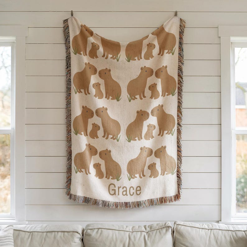 K&ouml;nnte beinhalten: Eine beige gewebte Decke mit einem sich wiederholenden Muster aus Capybaras und ihren Jungen. Die Decke hat einen Fransenrand und der Name "Grace" ist unten aufgedruckt. Die Capybaras sind in Braunt&ouml;nen gehalten, mit gr&uuml;nen Grasdetails.