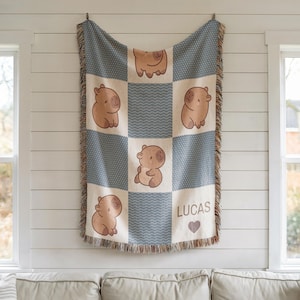 May include: A woven blanket with a patchwork design featuring light brown capybara illustrations on a cream and blue background. The blanket has a fringed edge and the name "LUCAS" with a heart symbol.