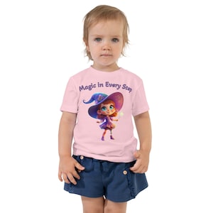 May include: A pink toddler t-shirt with a cartoon illustration of a girl wearing a purple witch hat and holding a wand. The text "Magic in Every Step" is printed on the shirt.