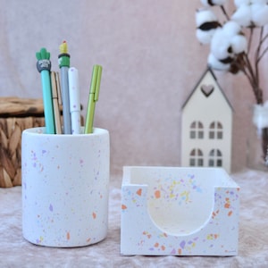 May include: White ceramic desk organizer set with a speckled pattern. The set includes a pen holder and a note holder. The pen holder has five pens inside, including a green pen with a cactus topper.