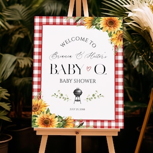 May include: A baby shower welcome sign on an easel. The sign has a red and white checkered border and features the text "Welcome to Brianna & Hector's Baby Q Baby Shower" with a barbecue grill graphic and sunflowers.