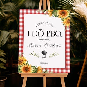 May include: A wedding welcome sign on an easel. The sign has a red and white checkered border and features the text "Welcome to the I Do BBQ" with the names Briana & Hector. Sunflowers and greenery decorate the corners.