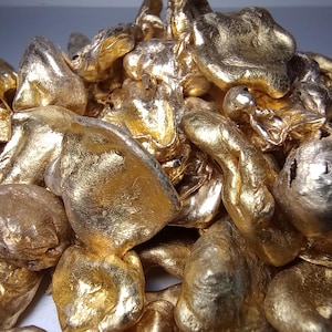 May include: A close-up of a pile of gold nuggets. The nuggets have a rough, textured surface and vary in size and shape. The lighting creates highlights and shadows, enhancing the metallic sheen.