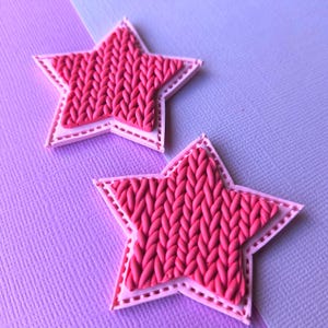May include: Two star-shaped ornaments featuring a bright pink centre with a raised, textured knit pattern. Each star is framed by a light pink border with a subtle stitched detail along the edges, creating a layered appearance for these items.