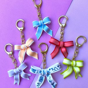 May include: Six decorative bow keyrings in pastel shades of blue, pink, peach, lilac, and green. Each bow is attached to a gold-coloured keyring with a clasp. The keyrings are displayed against a purple background.