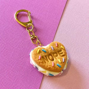May include: A gold-toned metal keyring attached to a heart-shaped cookie sandwich charm. The biscuit features the embossed text NOPE and is decorated with small, colourful sprinkles. A thick layer of white cream filling is visible between the biscuits.