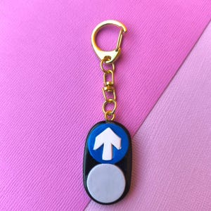 May include: A black and blue keyring with a white upward-pointing arrow and a white circle below. The keyring has a gold-coloured clasp and chain. The background is pink and purple.