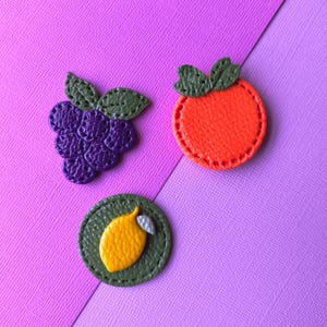 May include: Three fruit-shaped leather patches: a bunch of purple grapes, an orange persimmon, and a yellow lemon. Each patch has green leaves and is set against a pink and purple background. These patches are likely for crafts or embellishments.