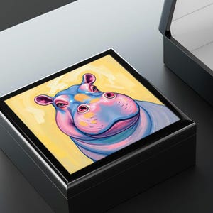 May include: A black wooden box with a hinged lid. The lid features a colorful painting of a hippopotamus with pink, blue, and yellow hues. The hippopotamus has a friendly expression and is looking directly at the viewer.