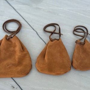 May include: Three brown suede drawstring pouches of varying sizes. The pouches have brown leather drawstrings and are arranged in a row from largest to smallest.