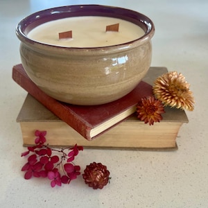 Op de afbeelding: Een bruine keramische kaars pot met een paarse rand en twee houten lonten. De kaars staat op een stapel boeken met gedroogde bloemen.