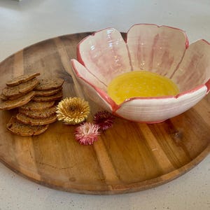 May include: A pink and white ceramic bowl shaped like a flower with a yellow centre. The bowl is sitting on a wooden tray with a stack of crackers.