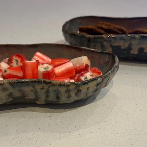 Puede incluir: Dos fuentes de cerámica hechas a mano. Una contiene caramelos rojos y blancos, la otra galletas. Las fuentes tienen un esmalte texturizado azul oscuro y marrón, y tienen forma ovalada.