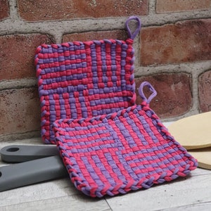 Könnte beinhalten: Zwei quadratische Topflappen aus gewebtem Stoff in Lila und Rosa mit einer Schlaufe. Die Topflappen sind aus weichem, gewebtem Stoff gefertigt und eignen sich perfekt zum Schutz der Hände vor heißen Oberflächen.