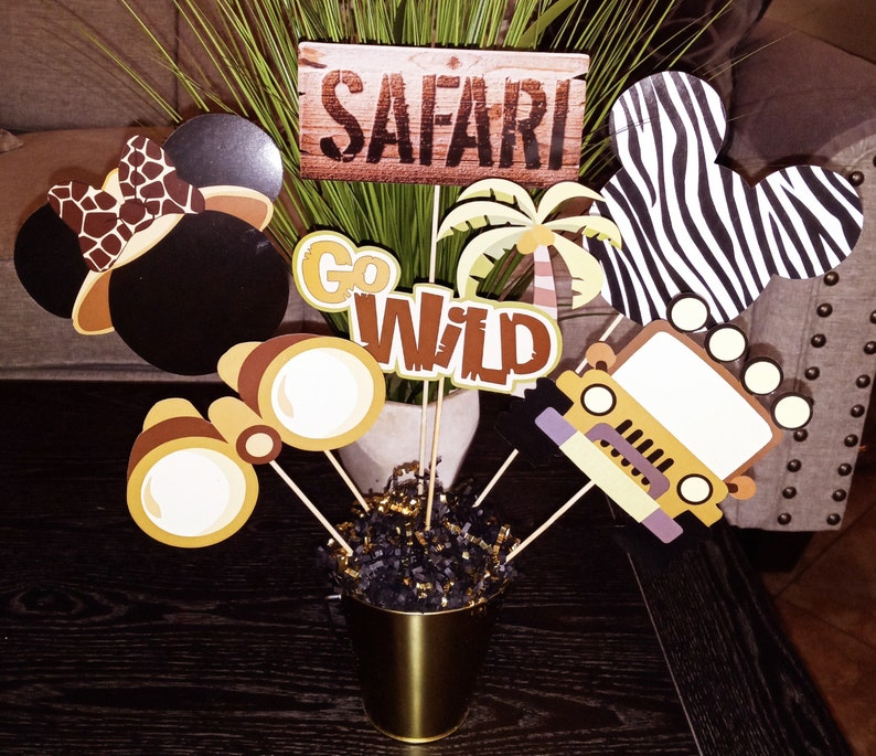 May include: A safari-themed centerpiece with a variety of decorations, including a pair of binoculars, a jeep, a palm tree, a zebra print Mickey Mouse ear, a brown Mickey Mouse ear with a leopard print bow, and a sign that says "Go Wild".