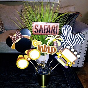 May include: A safari-themed centerpiece with a wooden sign that says "Safari", a green plant, a black and white zebra Mickey Mouse head, a yellow jeep, a pair of binoculars, and a brown and yellow Mickey Mouse head with a brown and yellow safari hat.