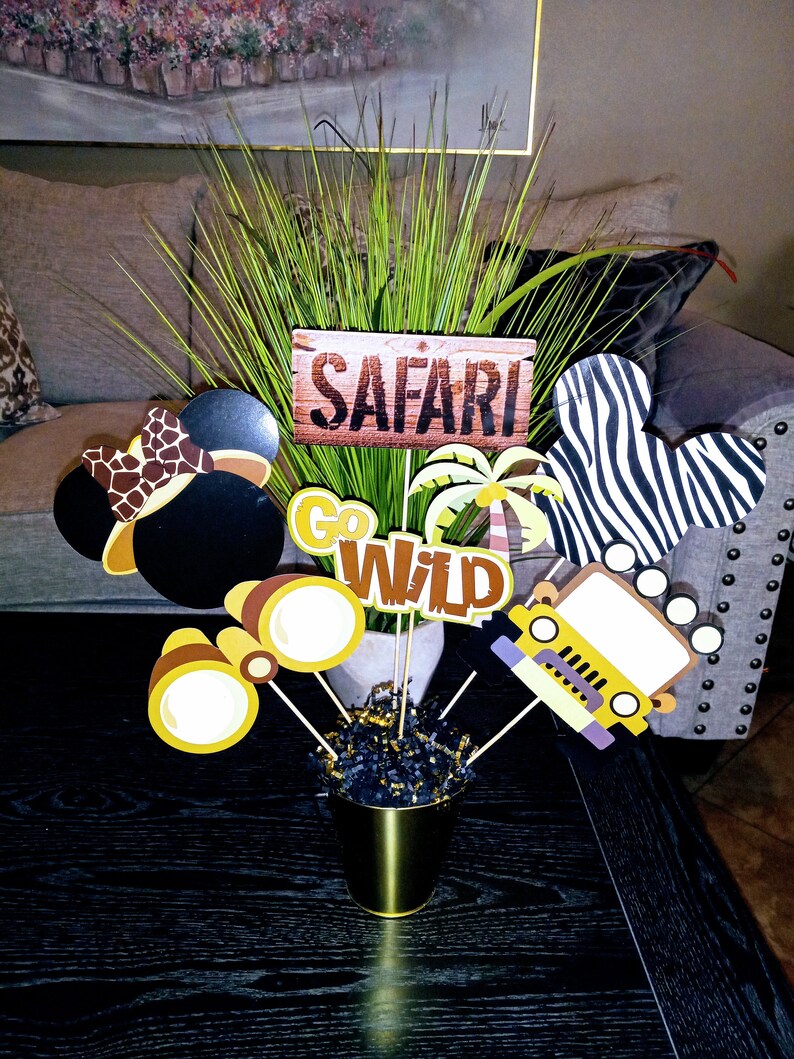 May include: A safari-themed centerpiece with a wooden sign that says "Safari", a green plant, a black and white zebra Mickey Mouse head, a yellow jeep, a pair of binoculars, and a brown and yellow Mickey Mouse head with a brown and yellow safari hat.