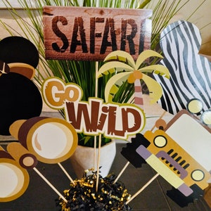 May include: A safari-themed centerpiece with a wooden sign that says "SAFARI", a palm tree, a jeep, binoculars, and a black and white zebra print. The centerpiece is in a gold vase with black and gold shredded paper.