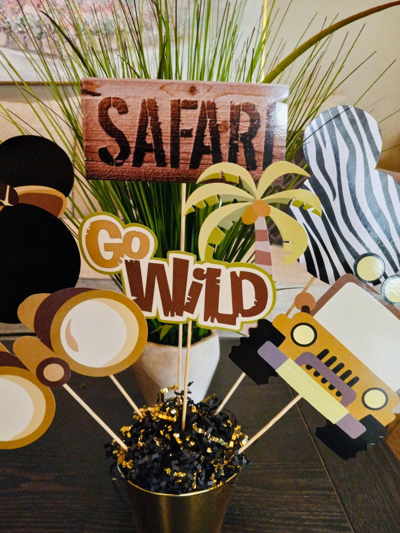 May include: A safari-themed centerpiece with a wooden sign that says "SAFARI", a palm tree, a jeep, binoculars, and a black and white zebra print. The centerpiece is in a gold vase with black and gold shredded paper.