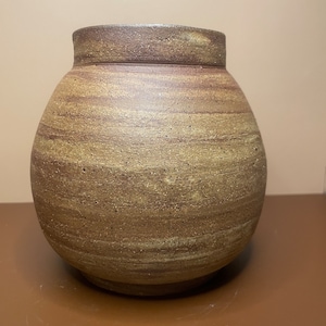 May include: A brown ceramic vase with a rounded body and a slightly flared rim. The vase has a speckled and streaked appearance, with shades of brown, tan, and beige.