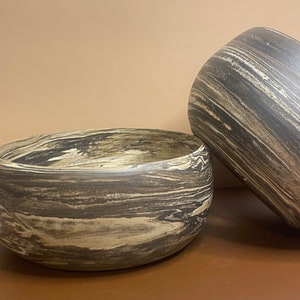 Puede incluir: Dos cuencos de cerámica con un diseño marmoleado en negro y marrón. Los cuencos son redondos y tienen un acabado liso y brillante.