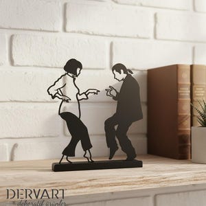 May include: A black metal silhouette sculpture depicting two figures in a dancing pose, mounted on a rectangular base. The design features a person with a bob haircut and a person with long hair, both captured in a mid-dance motion. The brand name DERVART dekoratif &uuml;r&uuml;nler is printed in the bottom left corner.