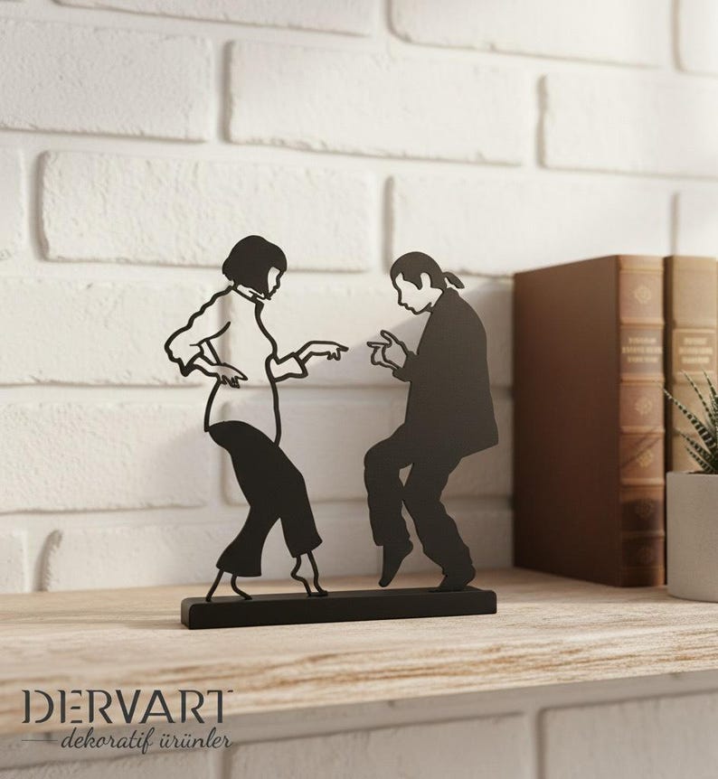 May include: A black metal silhouette sculpture depicting two figures in a dancing pose, mounted on a rectangular base. The design features a person with a bob haircut and a person with long hair, both captured in a mid-dance motion. The brand name DERVART dekoratif &uuml;r&uuml;nler is printed in the bottom left corner.