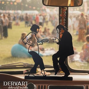 May include: A black metal silhouette sculpture depicting two figures in a dancing pose, mounted on a rectangular base. The design features the text DERVART dekoratif &uuml;r&uuml;nler at the bottom left. The object is positioned on a vehicle dashboard.