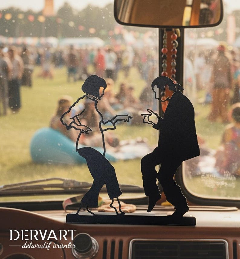 May include: A black metal silhouette sculpture depicting two figures in a dancing pose, mounted on a rectangular base. The design features the text DERVART dekoratif &uuml;r&uuml;nler at the bottom left. The object is positioned on a vehicle dashboard.