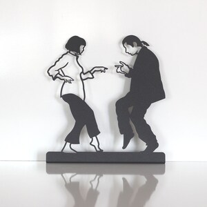 May include: A black metal wall art piece depicting two figures in a dancing pose. The silhouette shows a person with a bob haircut wearing a shirt and pants, and another person with long hair in a ponytail wearing a suit, both pointing fingers.