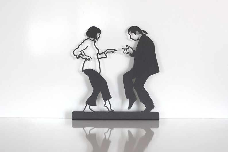 May include: A black metal wall art piece depicting two figures in a dancing pose. The silhouette shows a person with a bob haircut wearing a shirt and pants, and another person with long hair in a ponytail wearing a suit, both pointing fingers.