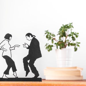 May include: A black metal silhouette sculpture depicting two figures in a dancing pose. The figure on the left has short hair and wears a long-sleeved top and cropped pants. The figure on the right has long hair tied back and wears a suit jacket and trousers.