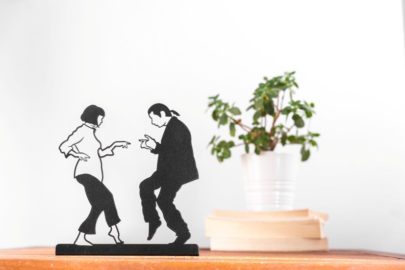 May include: A black metal silhouette sculpture depicting two figures in a dancing pose. The figure on the left has short hair and wears a long-sleeved top and cropped pants. The figure on the right has long hair tied back and wears a suit jacket and trousers.