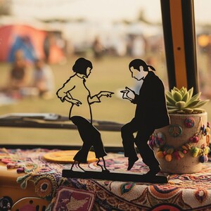 May include: A black metal silhouette sculpture depicting two figures in a dancing pose, mounted on a rectangular base. The design features a person with a bob haircut and a person with a ponytail, both dressed in shirts and trousers, mid-dance.