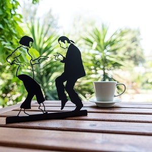 May include: A black metal silhouette sculpture depicting two figures dancing, mounted on a rectangular base. Beside the sculpture sits a white ceramic coffee cup and saucer. The figures are captured in a dynamic pose with arms extended.